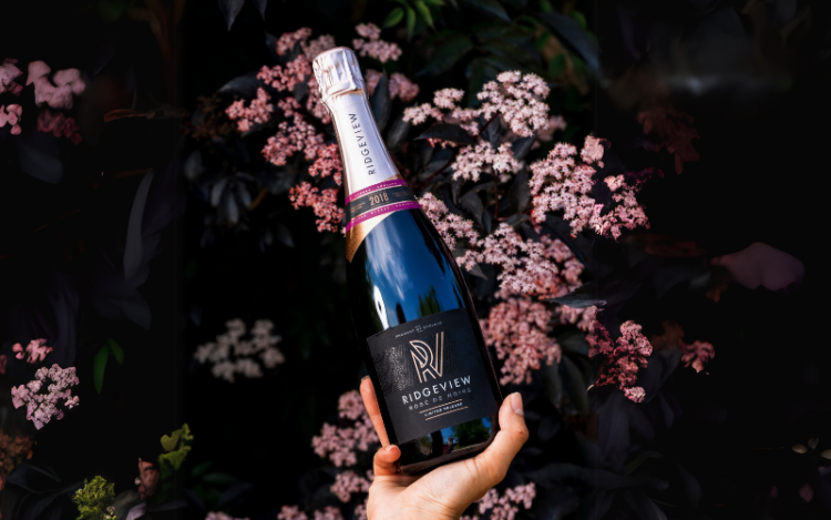 A hand holding a bottle of Rosé de Noirs above pink flowers outside