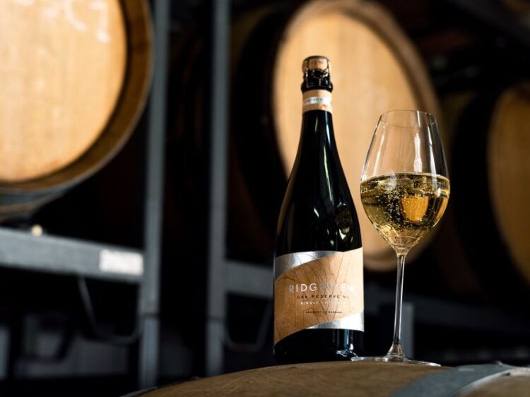 A bottle of Ridgeview Oak Reserve and a full glass, on an oak barrel with oak barrels on racks in the background in the Ridgeview Winery.