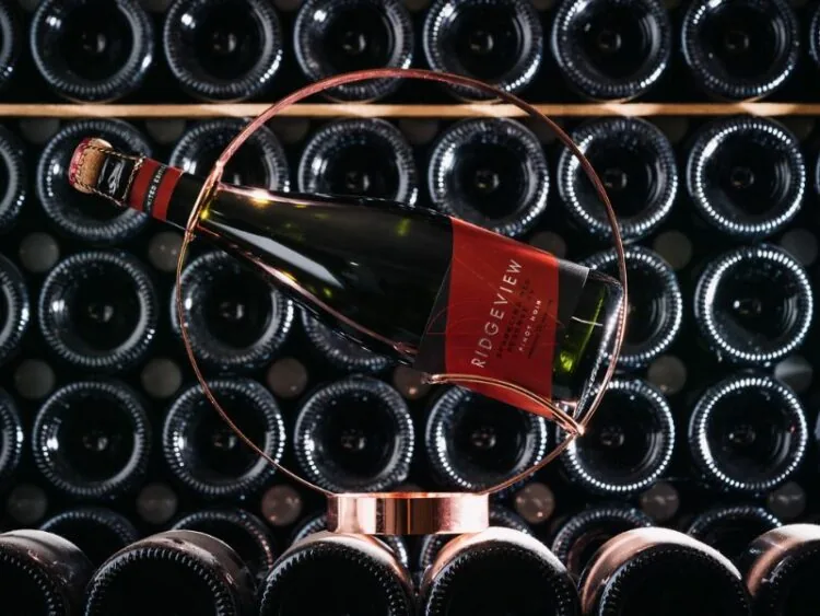 A bottle of Ridgeview's Sparkling Red Reserve horizontal in an ornamental stand, in front of bottles on a wine rack