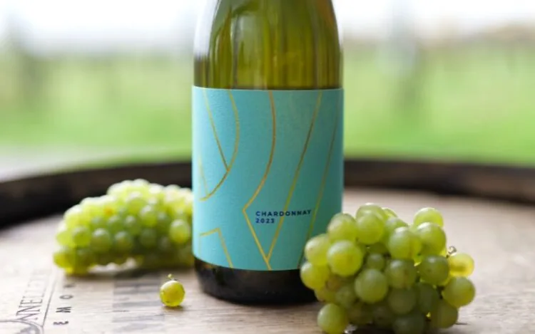 A bottle of Ridgeview's still Chardonnay with a bright blue label, on an oak barrel with Chardonnay grapes and the vineyard in the background.